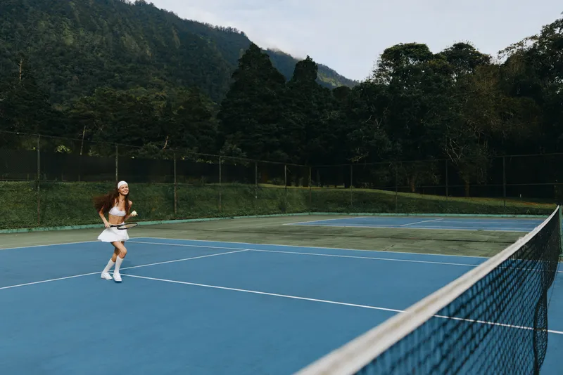 Young female tennis player practicing on outdoor court surrounded by greenery, expressing focus and determination while dressed in sporty attire
