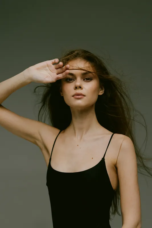 Young woman posing with hand on forehead wearing black dress against plain background