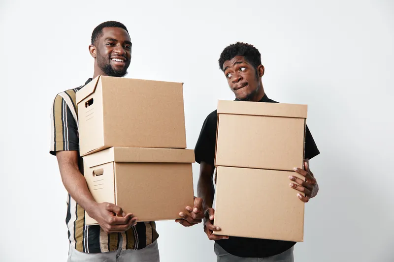 Two black men carrying cardboard boxes for moving or delivery, smiling and standing against white background in casual clothes with dark skin and happy expressions.
