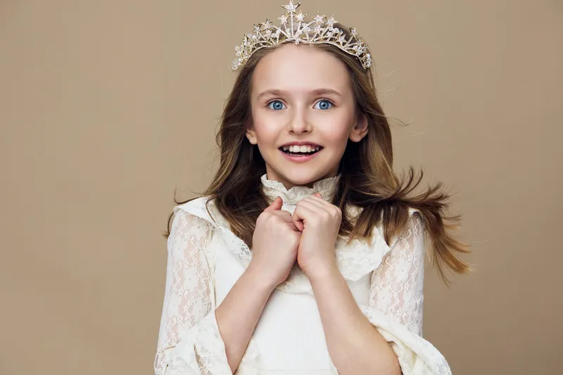 A joyful young girl with a crown, dressed in a vintage white dress, smiling brightly against a neutral beige background, representing innocence and childhood wonder