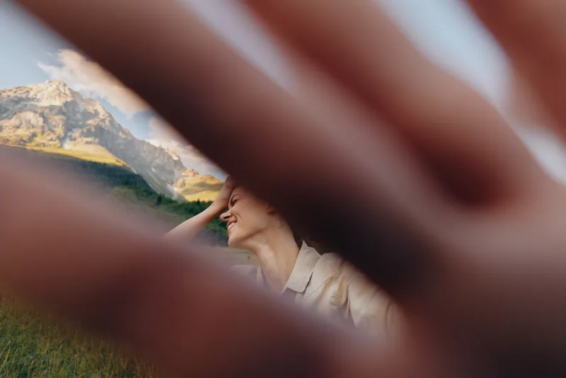 Young woman smiling outdoors with hand partially covering camera, mountain landscape in background, natural light, concept of lifestyle and happiness.