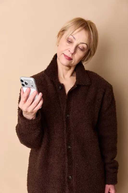 Elderly woman holding smartphone indoors, warm beige background, thoughtful gaze while texting, relaxed senior portrait about communication and technology use, calm cozy coat