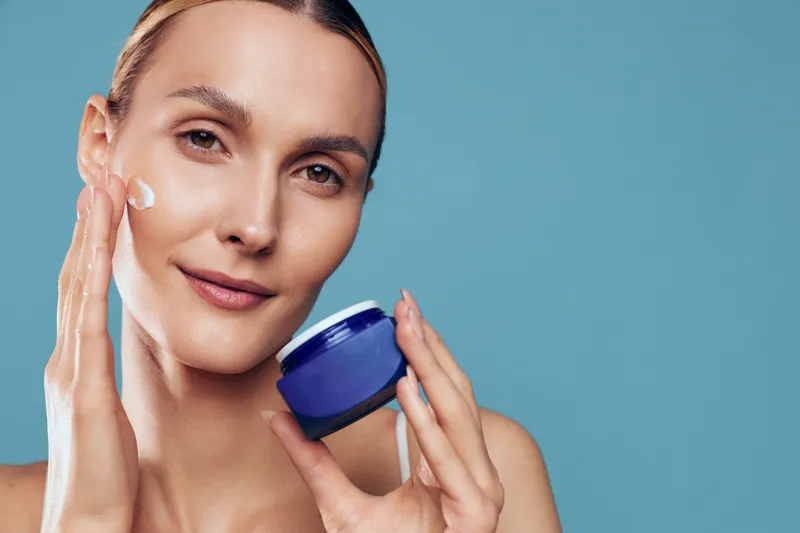 Young woman applies moisturizing cream on face holds blue jar skincare product demonstrates daily beauty routine isolated on blue background with natural healthy skin and soft expression