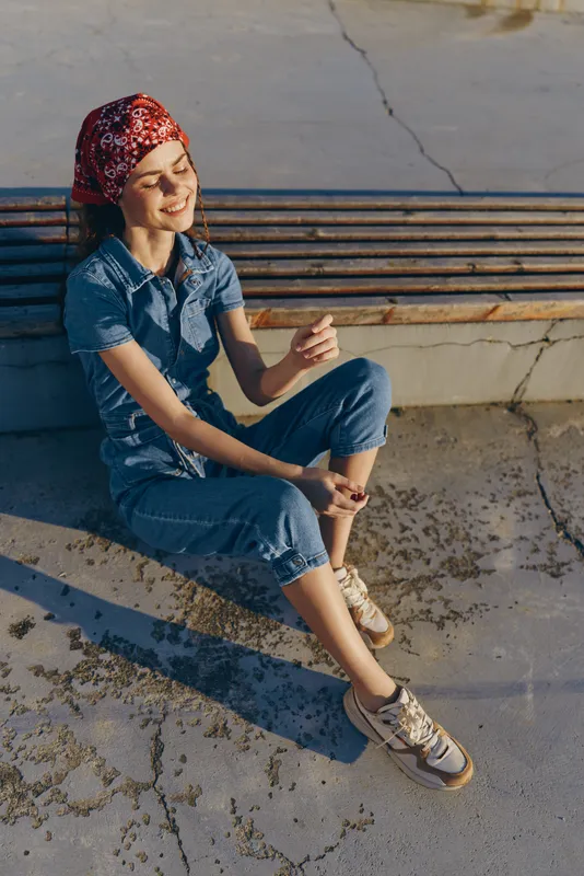 Lifestyle woman in modern boho-western denim street style sitting casually outdoors with a relaxed smile, wearing a red bandana and sneakers, film color effect.