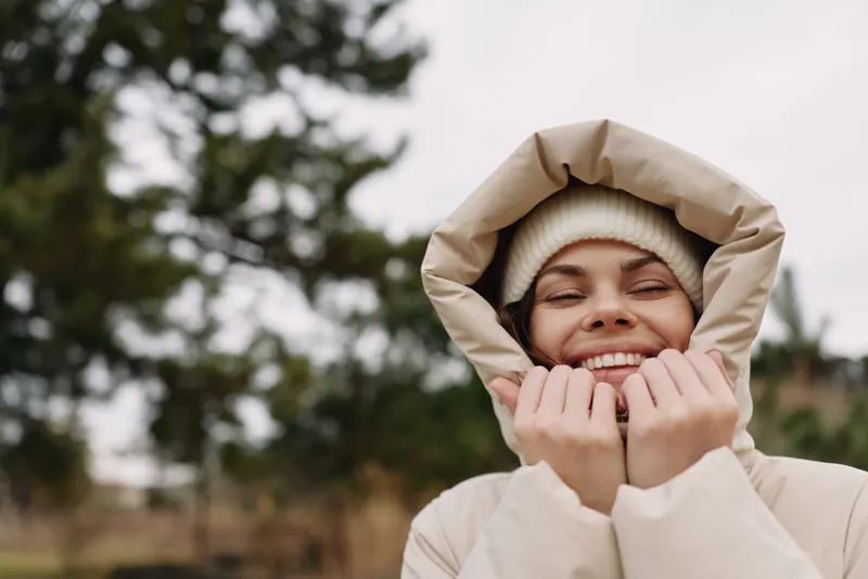 Woman in a warm hooded jacket and beanie smiling at the camera in a chilly outdoor setting, soft natural light, trees in the background, cozy winter mood