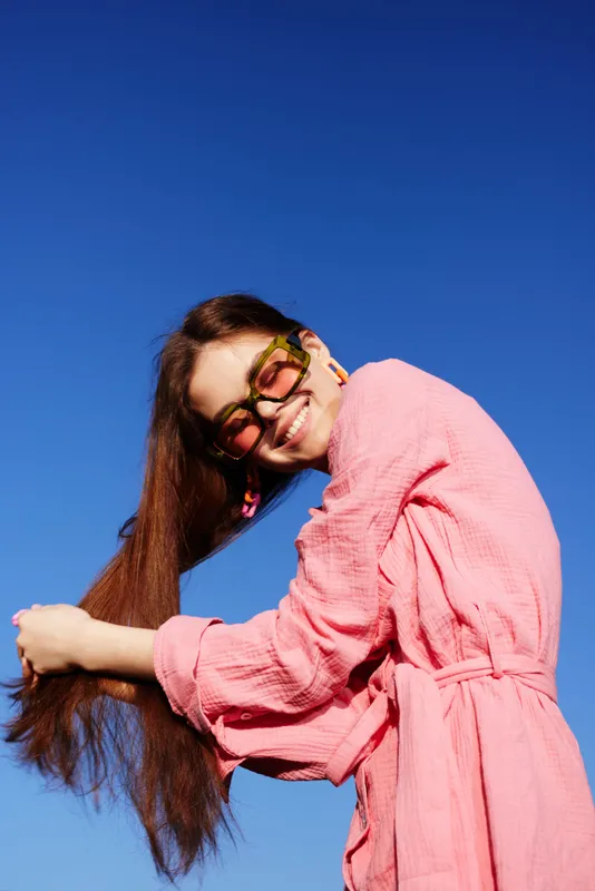 Happy young woman smiling and playing with her long hair against a clear blue sky, wearing a stylish pink dress and sunglasses Perfect for lifestyle themes