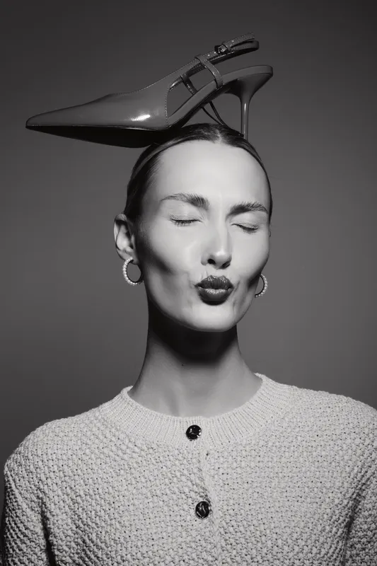 A white woman with pale skin and closed eyes balances a high heel shoe on her head. She wears hoop earrings and a textured sweater in a black and white studio portrait. Concept of fashion and balance.
