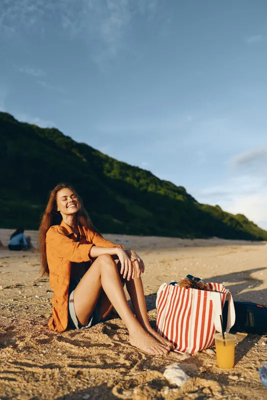 Woman on beach in summer, sitting on sand near striped bag, smiling and relaxing, wearing orange dress, sunshine and ocean in background, nature travel mood, soft light, breezy air