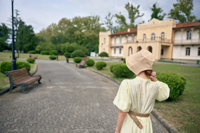 Woman in a vintage dress and straw hat walks along a park path toward a grand estate, creating a serene outdoor scene with timeless charm today.