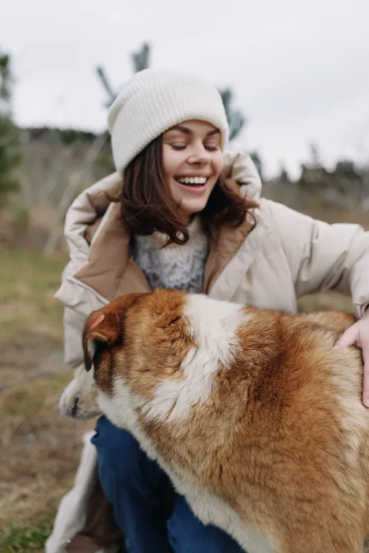 Woman in a knit beanie and warm coat smiles while hugging a fluffy dog outdoors on a crisp day, capturing a cozy moment of companionship and gentle joy in nature