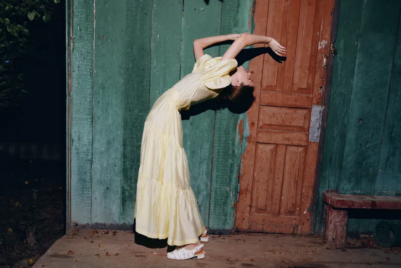 Woman in a pale yellow dress arches backward beside a weathered wooden door in a dim outdoor setting, creating a graceful, moody artistic moment with cinematic atmosphere