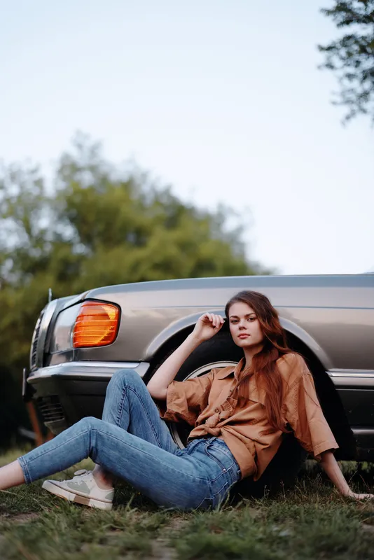 Stylish young woman with long hair sitting next to a vintage car in a natural setting, wearing a brown top and jeans, evoking a carefree summer vibe.