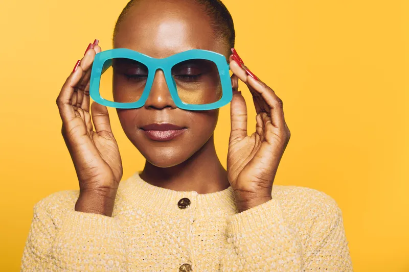 Close-up of a confident black woman with dark skin holding oversized blue glasses near her face. She wears a yellow knitted sweater against a bright yellow background. Concept of style and fashion.