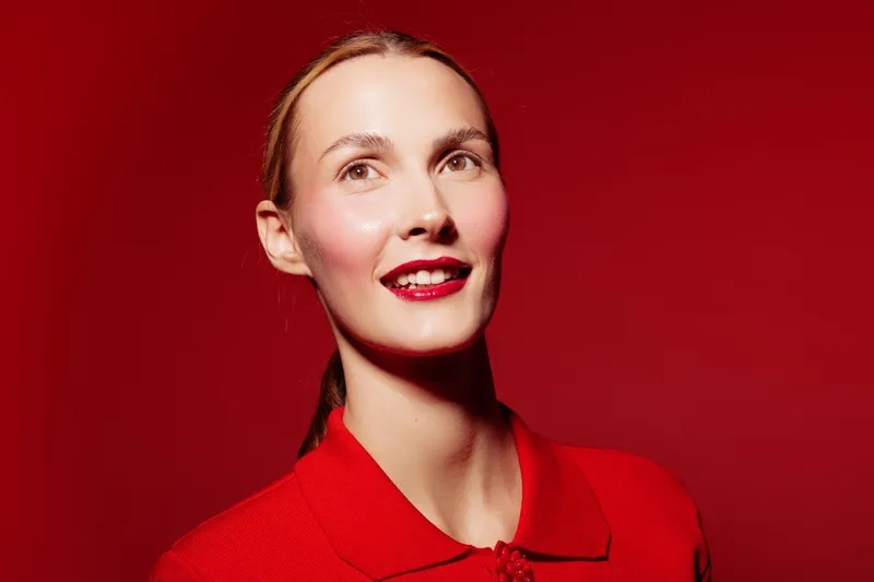 Smiling woman with red lipstick and red shirt poses against red background. Studio portrait with bright lighting highlights natural skin and confident expression. Concept of beauty and fashion.