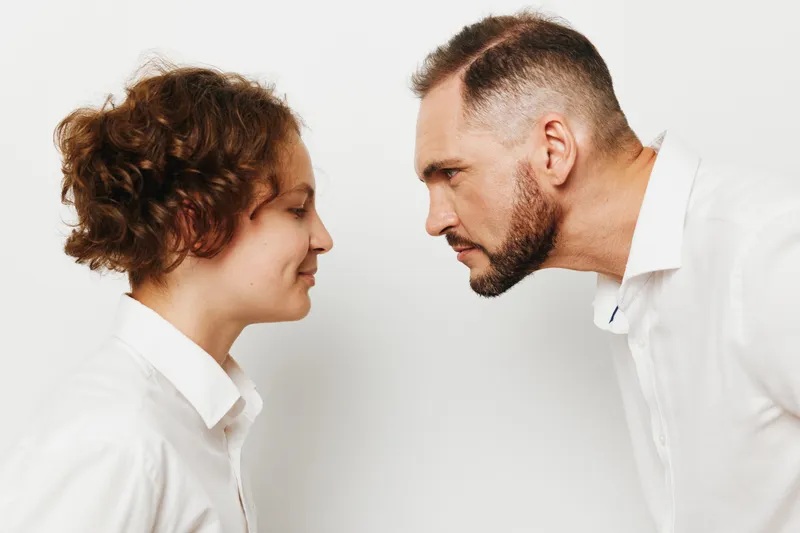 A focused professional man in a white shirt stands against a solid colored background, sharing a calm, confident moment with a colleague, conveying strong business presence.