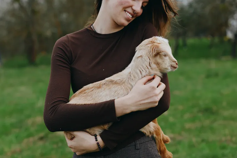 A woman in a brown sweater holds a pale goat kid in a grassy field, conveying tender countryside lifestyle, animal companionship, and calm wellness. Soft natural light creates a warm, affectionate
