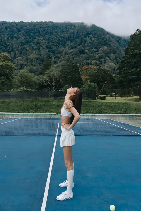 Young female tennis player stretching on a court, wearing a white sports outfit, conveying joy and confidence against a lush green mountain backdrop