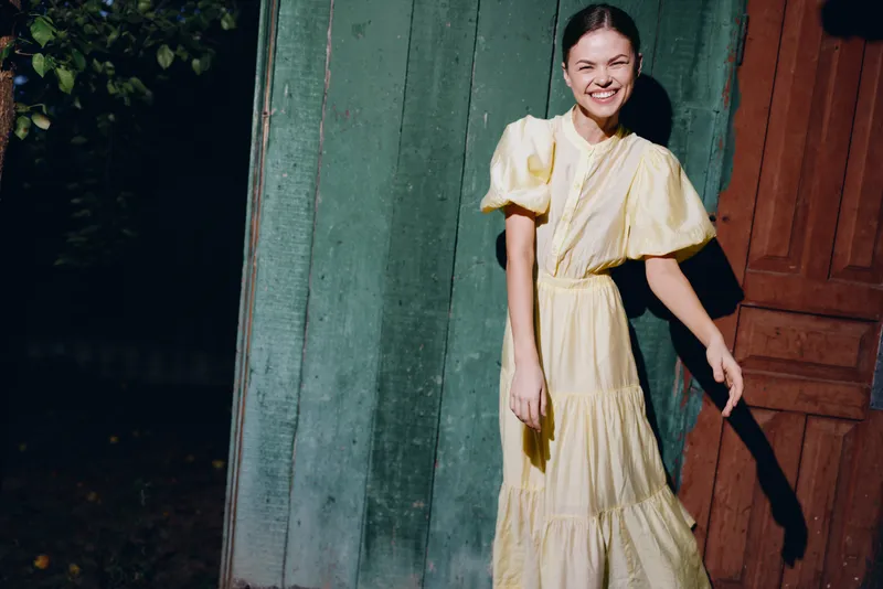 A smiling woman in a yellow dress stands outdoors beside a doorway, bathed in warm sunlight, showcasing casual fashion and a joyful candid moment in a sunny setting.