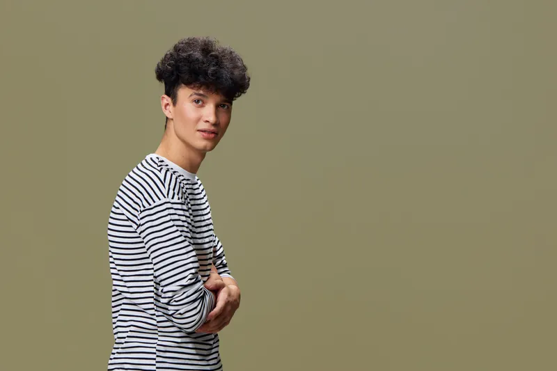 young man with curly hair wearing striped shirt standing confidently against plain green background, casual style and calm expression in studio setting