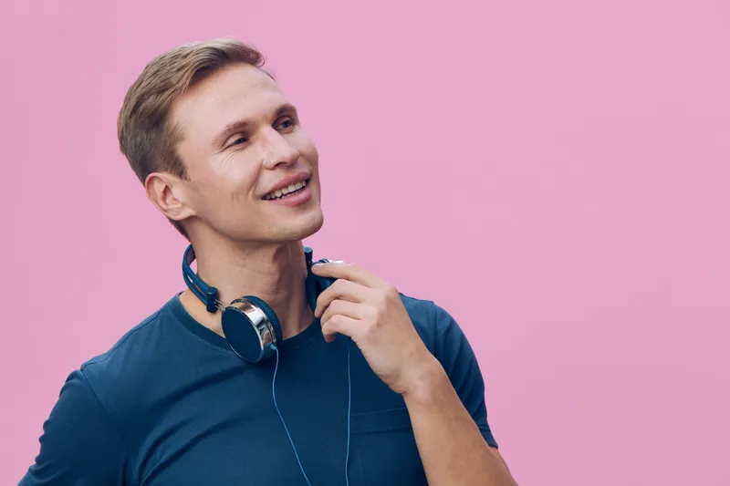 Happy young man with short blond hair in casual blue t-shirt poses with headphones around neck, smiling and looking slightly upwards on pink background. Studio portrait of cheerful male personality fo