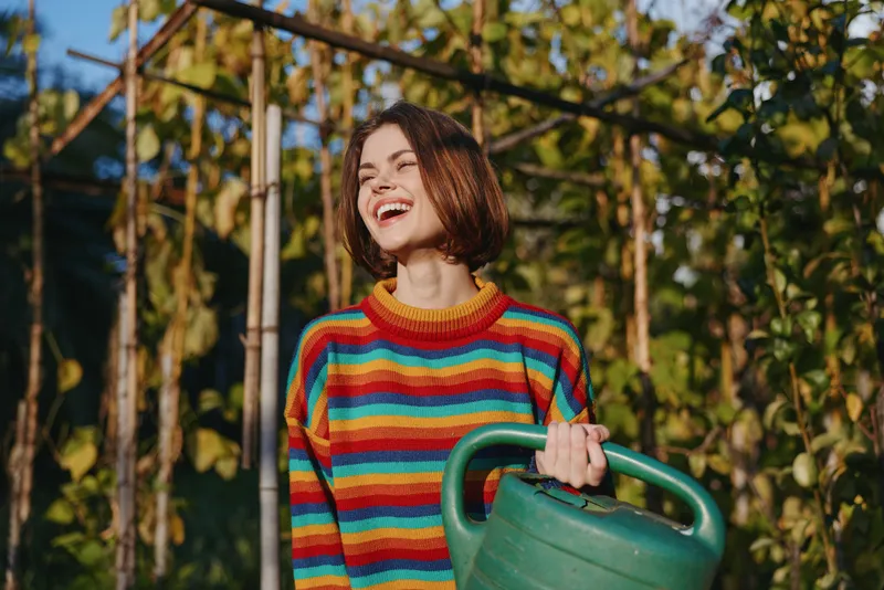 Young woman mid 20s in a striped sweater holding a watering can in a sunny garden by a wooden trellis, smiling and laughing in a casual outfit for lifestyle gardening.