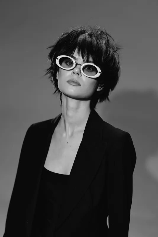 Stylish woman wearing white round sunglasses and black outfit in black and white portrait with short hair.