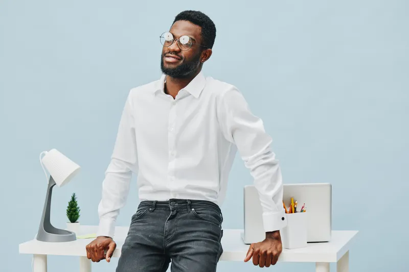Confident young Black man in white shirt and black jeans standing in modern office with smile, glasses, and relaxed pose, holding papers and leaning on desk. Studio shot, professional business style, 