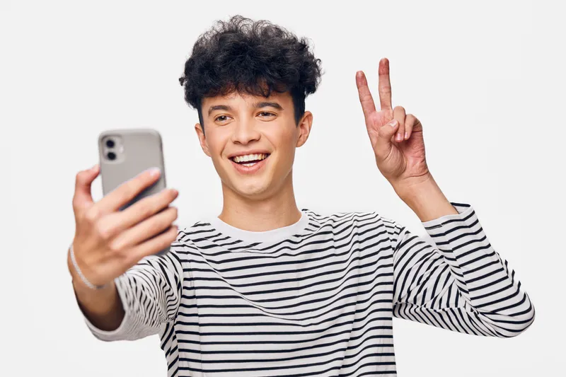 smiling young man wearing a striped shirt taking a selfie with peace sign against a white background, happy expression, casual style, isolated portrait