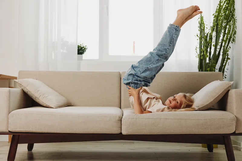 Happy child in casual clothes lies on beige sofa with legs raised in bright living room. Cozy home interior with natural light and green plant. Concept of playful childhood and comfort.