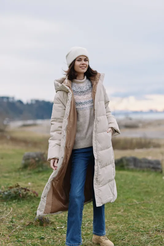 Female model wearing a long beige winter coat over a knit sweater and beanie, standing outdoors on a grassy countryside with a soft sky.