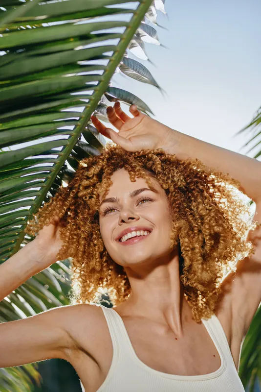 smiling woman with curly hair enjoying sunny day outdoors under palm leaves, natural light, relaxed mood, summer vibe, bright blue sky background