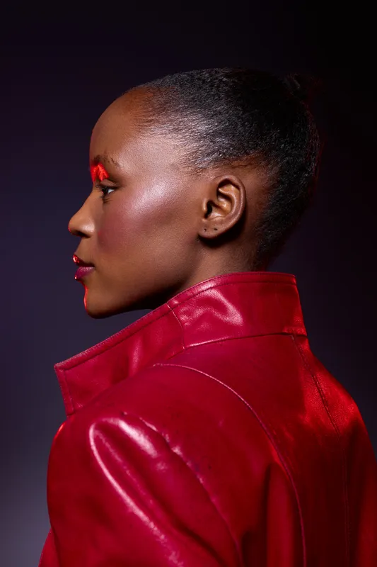 Profile of a dark skinned African woman wearing a red leather jacket and red eyeshadow. Studio portrait with dramatic lighting and smooth skin texture. Fashion and beauty concept.