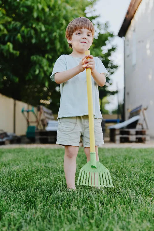 Young boy holding a rake in a sunny backyard, displaying curiosity and joy while engaging in outdoor activities among green grass and trees.