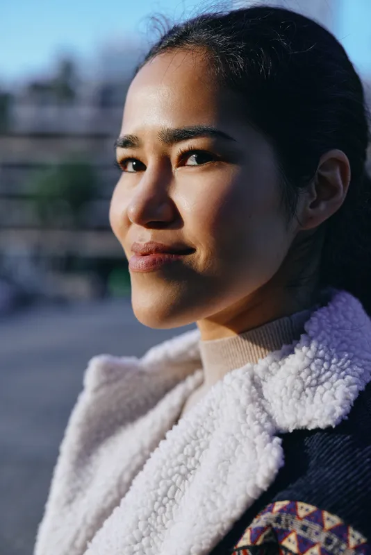 Closeup of smiling woman in outdoor setting with sunlight, denim jacket and shearling collar, calm expression, urban background, natural light, texture, and subtle warmth