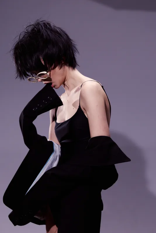 Young woman in black dress posing with messy hair and glasses against a gray background in modern fashion style