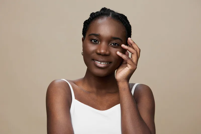 Portrait of a smiling Black woman with braided hair, dressed in a white top, against a soft beige background, conveying confidence and warmth
