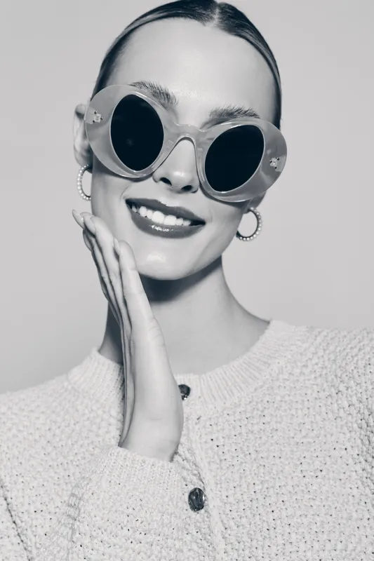 Young white woman with pale skin and sunglasses smiles, wearing hoop earrings and textured sweater in studio portrait.