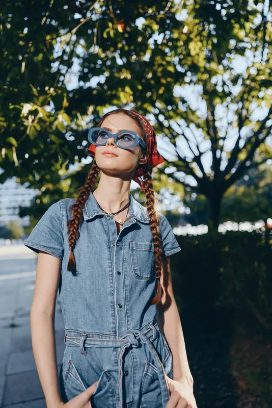Lifestyle woman wearing modern boho-western denim street style with sunglasses, standing confidently outdoors under green trees in warm natural light, film color effect.