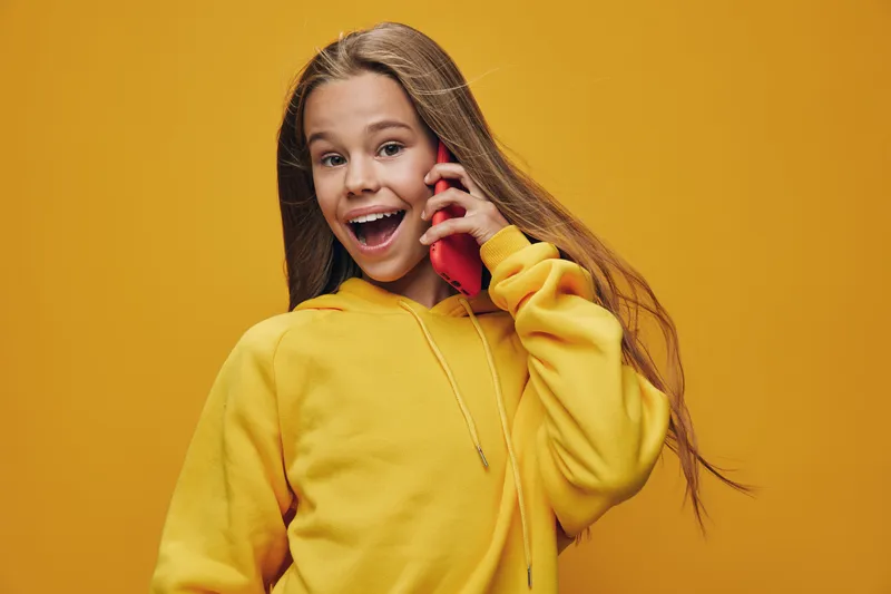 Girl wearing yellow hoodie smiling and talking on red phone with long hair against yellow background in studio. Happy teenager with casual style using mobile communication device.