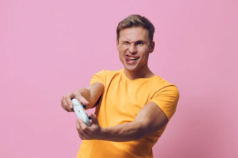 Playful young man with short brown hair wearing yellow t-shirt making funny face while holding game controller isolated on pink background studio portrait. People lifestyle concept