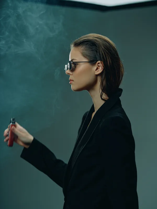 Stylish woman in sunglasses vaping with smoke on dark background, wearing black jacket, modern and confident atmosphere.