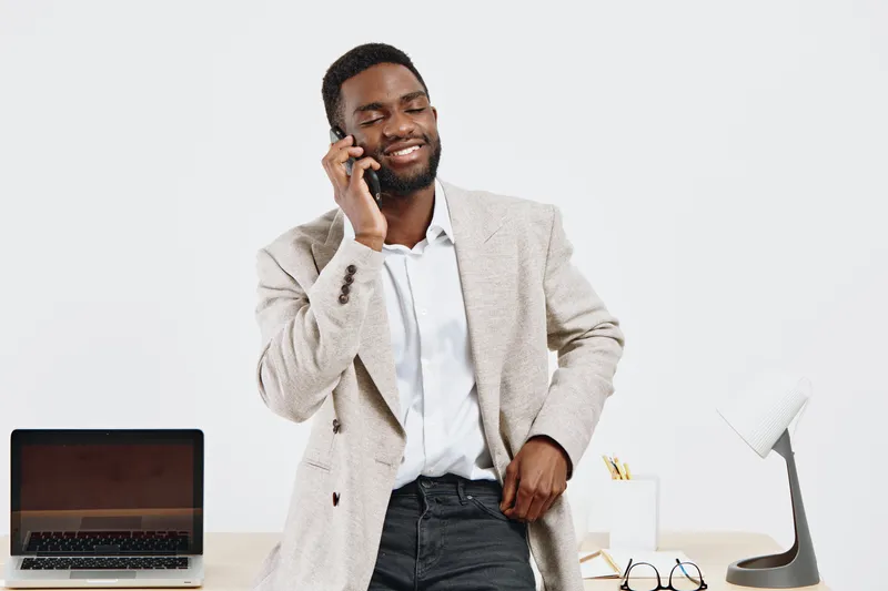 Confident smiling man talking on mobile phone while standing at desk in office, wearing smart casual suit, with laptop, glasses, and workspace accessories, modern professional workspace environment. P