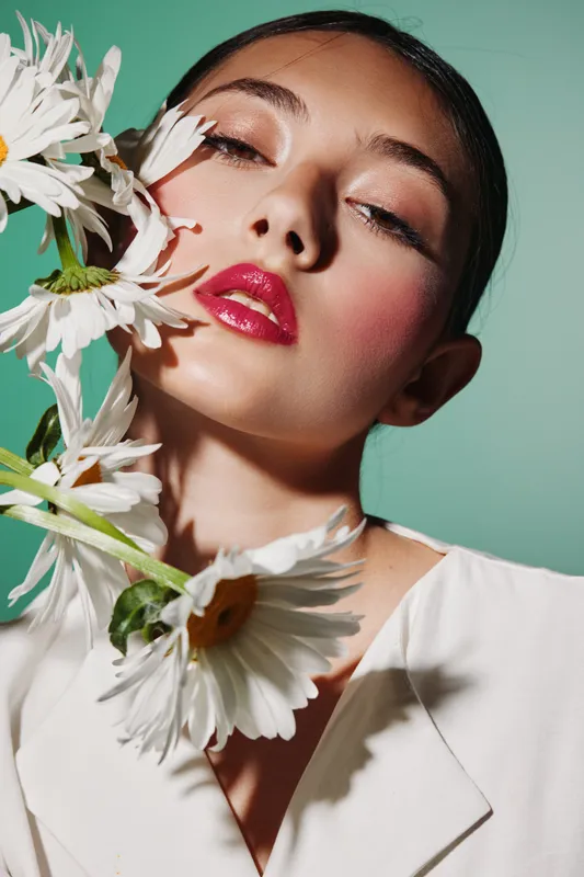 Close-up of a young woman with bright pink lipstick and creative pink eyeshadow surrounded by white daisies on a mint background. Concept of fashion, beauty, and creative makeup.