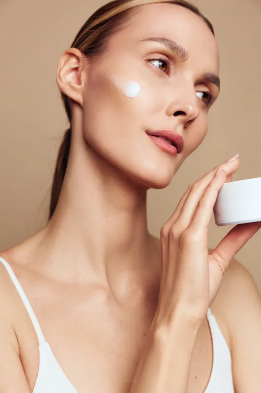 Young woman applies moisturizing cream on face with smooth skin holds jar of skincare product looks away with natural makeup and healthy glowing complexion isolated on beige background