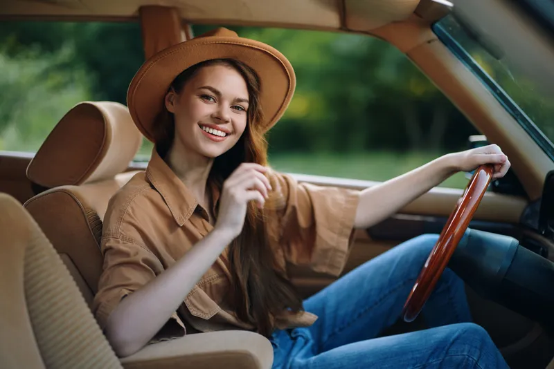 Young woman in a stylish hat smiling joyfully inside a vintage car, dressed in a casual beige outfit, evoking a mood of happiness in a natural green setting.