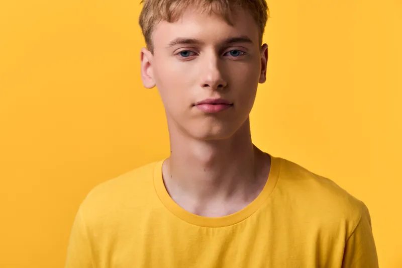 Young man in a yellow shirt stands against a yellow background with soft studio lighting, a calm expression, clean composition, and casual fashion for lifestyle use