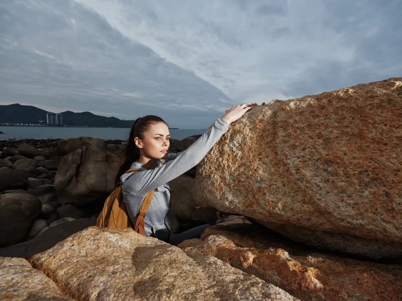 Young woman exploring rocky coast, wearing a backpack and a sporty outfit, showcasing determination and adventure against a cloudy background