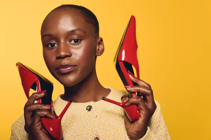 Confident dark skinned African woman holds red high heels near face on yellow background. Concept for fashion, style, beauty, and footwear with vibrant colors and confident expression.