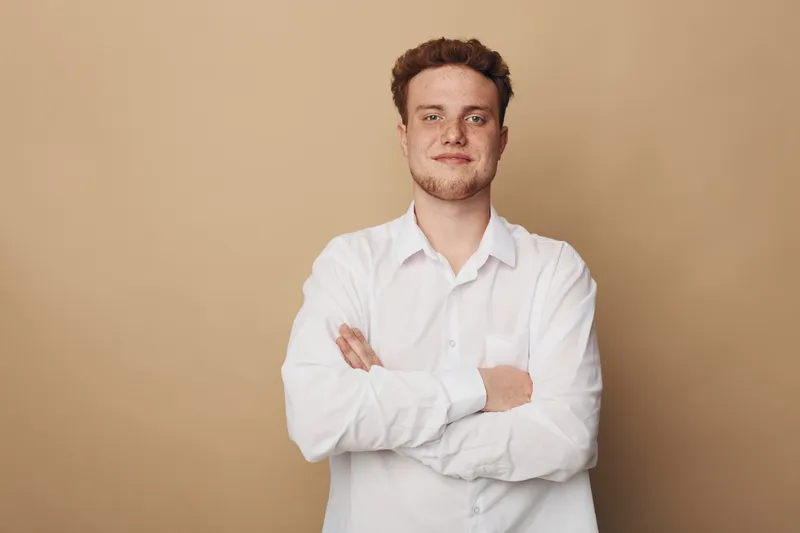 Confident young man with curly hair wearing white shirt stands with arms crossed against beige background. Portrait of casual male model with relaxed and self-assured expression.