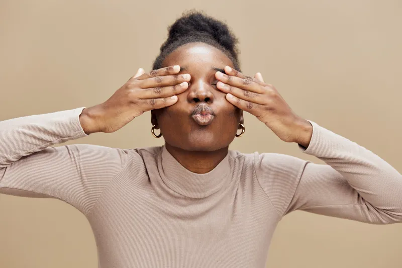 Happy African woman making playful faces while covering her eyes, wearing a beige turtleneck sweater, against a warm neutral background